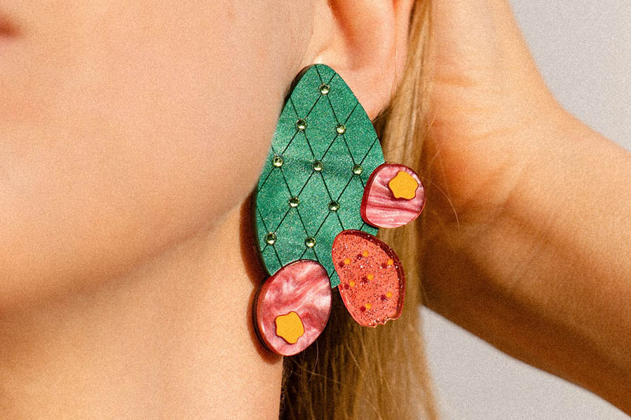 Close-up of a persons ear wearing a large, red, flower-shaped earring with green and black details, paired with a beige knit sweater.