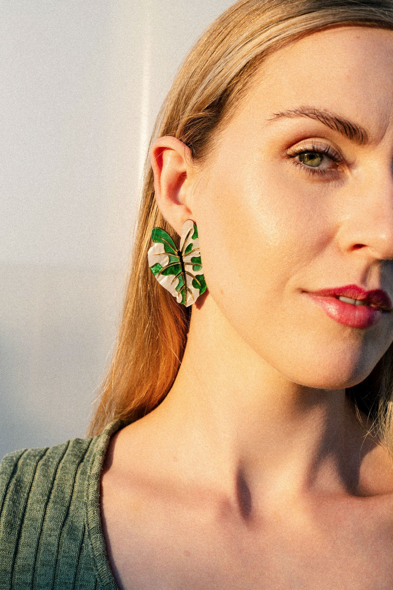 A woman with blonde hair wears a green ribbed top and a large Alocasia Earrings in a green and beige pattern. She is facing the camera in soft, natural light, showcasing the beauty of this hand-crafted jewelry.