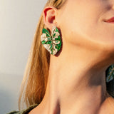 A close-up of a person wearing a large, green and white Alocasia Earrings, designed to resemble the intricate shape of leaves. The person’s head is tilted back, highlighting the hand-crafted jewelry and their neck. They have long blonde hair and are wearing a green top.