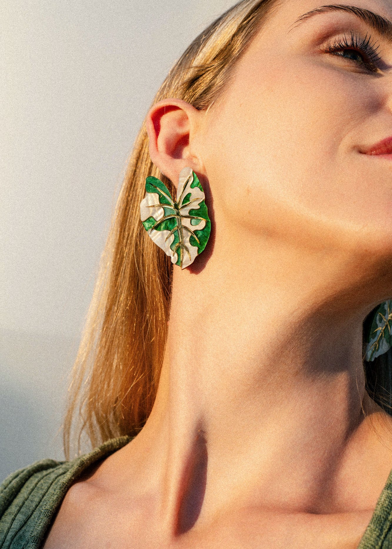 A close-up of a person wearing a large, green and white Alocasia Earrings, designed to resemble the intricate shape of leaves. The person’s head is tilted back, highlighting the hand-crafted jewelry and their neck. They have long blonde hair and are wearing a green top.