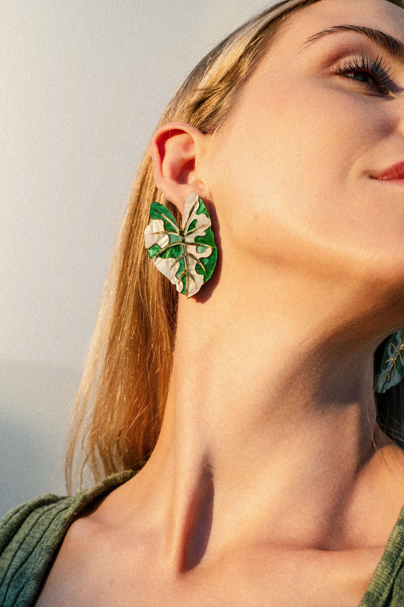 A close-up of a person wearing a large, green and white Alocasia Earrings, designed to resemble the intricate shape of leaves. The person’s head is tilted back, highlighting the hand-crafted jewelry and their neck. They have long blonde hair and are wearing a green top.
