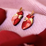 Two red Anatomical Heart Earrings with gold accents rest on a soft, pink textured fabric, surrounded by red rose petals—a unique style statement for acrylic earrings lovers.