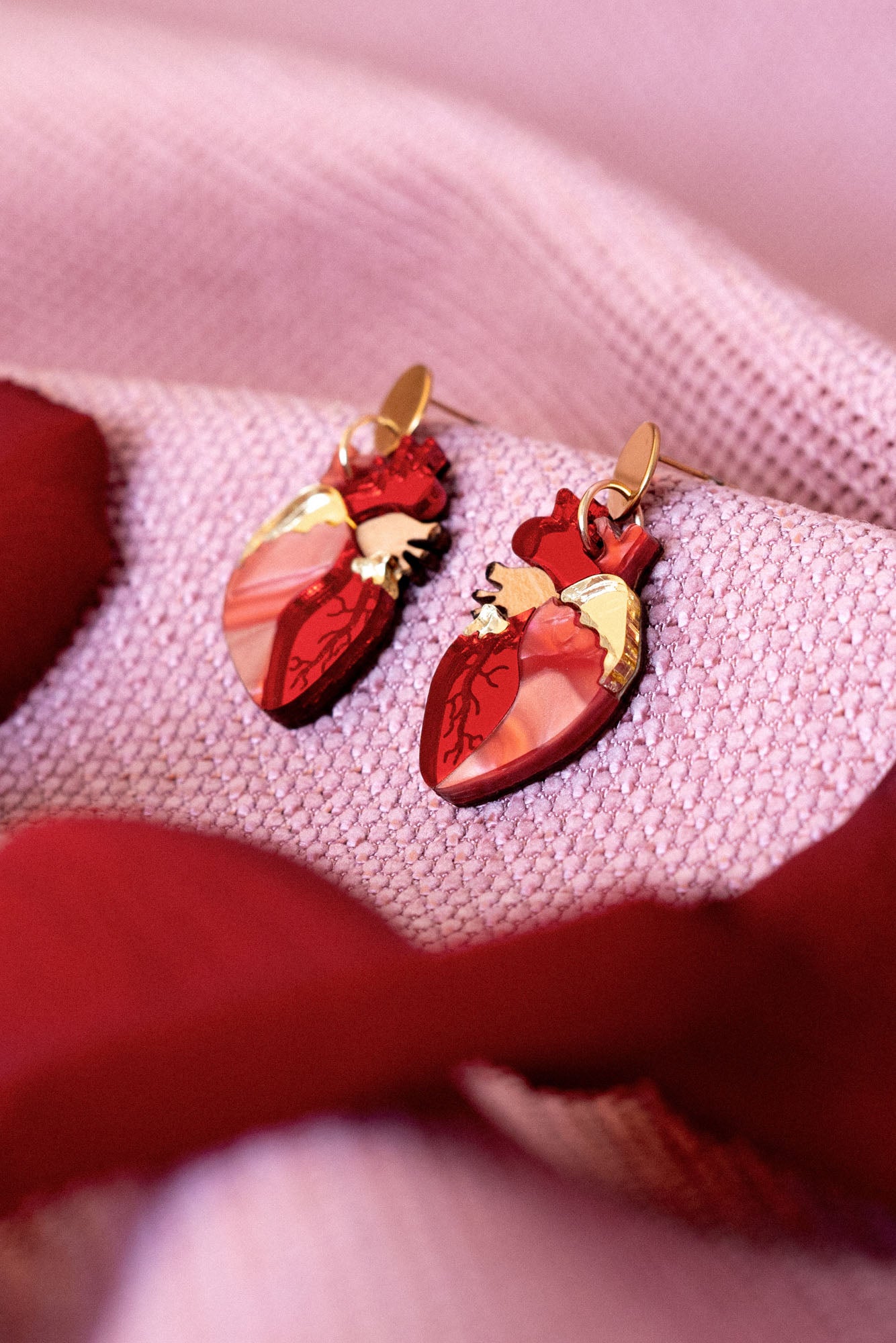 Two red Anatomical Heart Earrings with gold accents rest on a soft, pink textured fabric, surrounded by red rose petals—a unique style statement for acrylic earrings lovers.