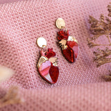 A pair of red and gold Anatomical Heart Earrings rest on textured pink fabric, with dried flowers nearby—acrylic earrings perfect for expressing your unique style.