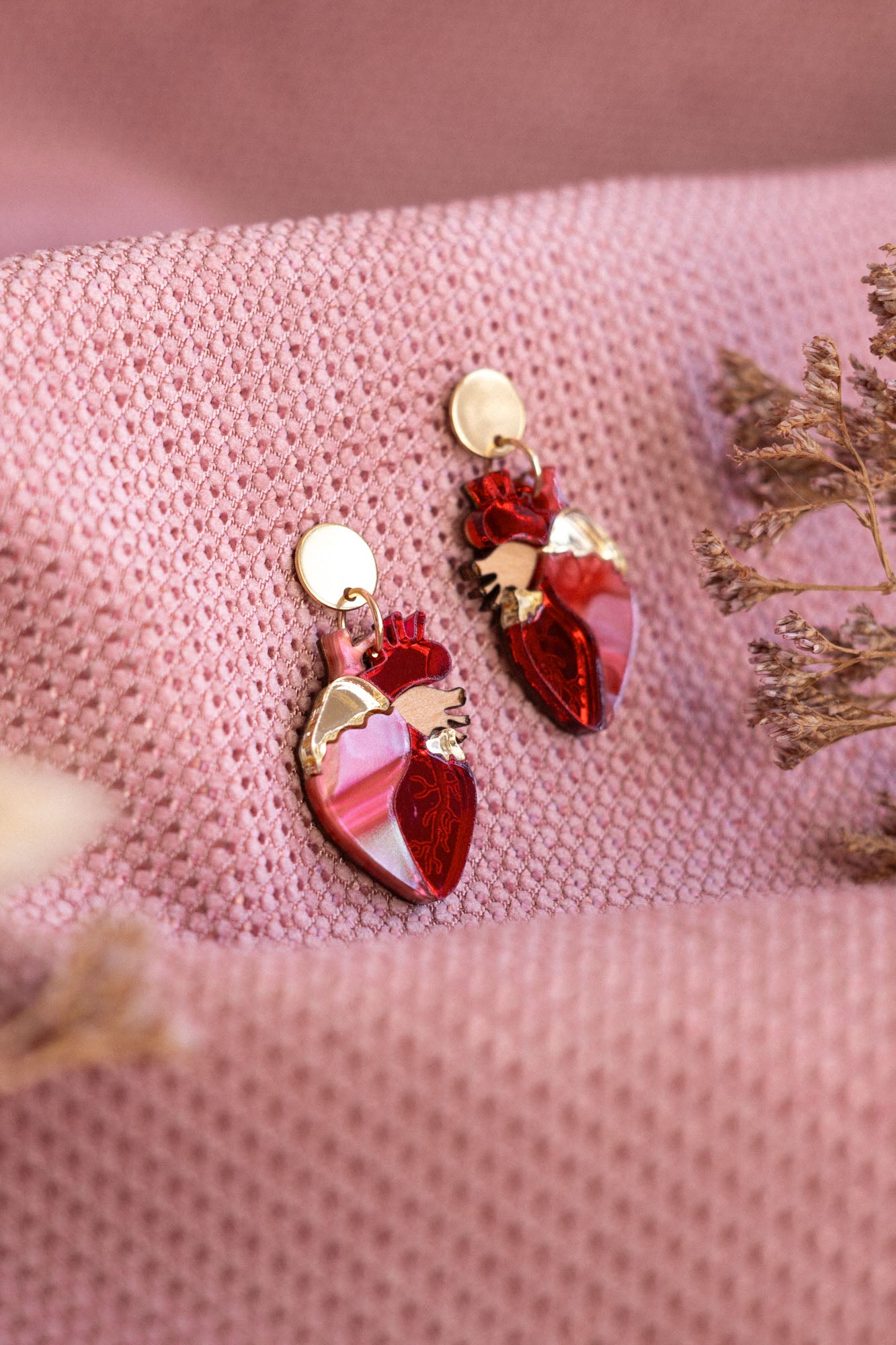 A pair of red and gold Anatomical Heart Earrings rest on textured pink fabric, with dried flowers nearby—acrylic earrings perfect for expressing your unique style.