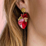 Close-up of a woman’s ear wearing unique style Anatomical Heart Earrings in red and pink tones with gold accents; her light brown hair is tucked behind her ear against a pink background, highlighting these striking acrylic earrings.