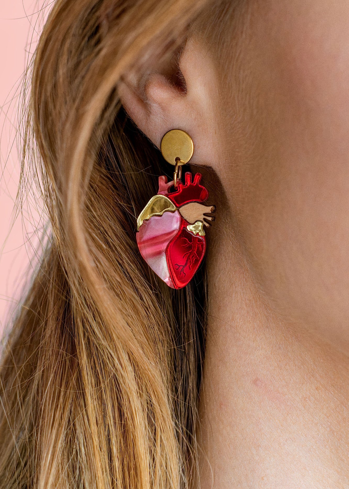 Close-up of a woman’s ear wearing unique style Anatomical Heart Earrings in red and pink tones with gold accents; her light brown hair is tucked behind her ear against a pink background, highlighting these striking acrylic earrings.