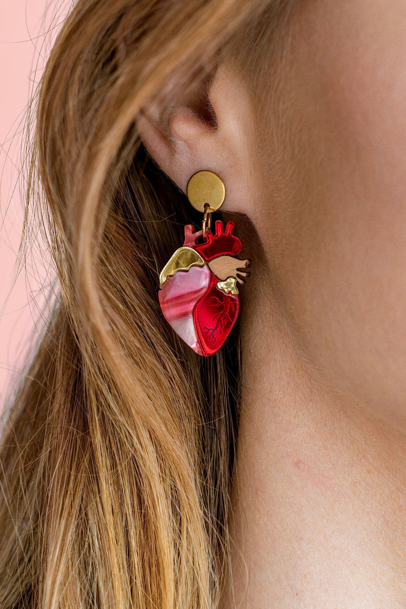 Close-up of a woman’s ear wearing unique style Anatomical Heart Earrings in red and pink tones with gold accents; her light brown hair is tucked behind her ear against a pink background, highlighting these striking acrylic earrings.