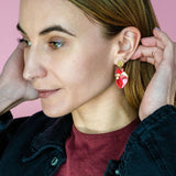 A woman with light brown hair wears a maroon shirt and black jacket, adjusting her unique acrylic earrings. She stands in front of a pink background, looking to the side with a slight smile.