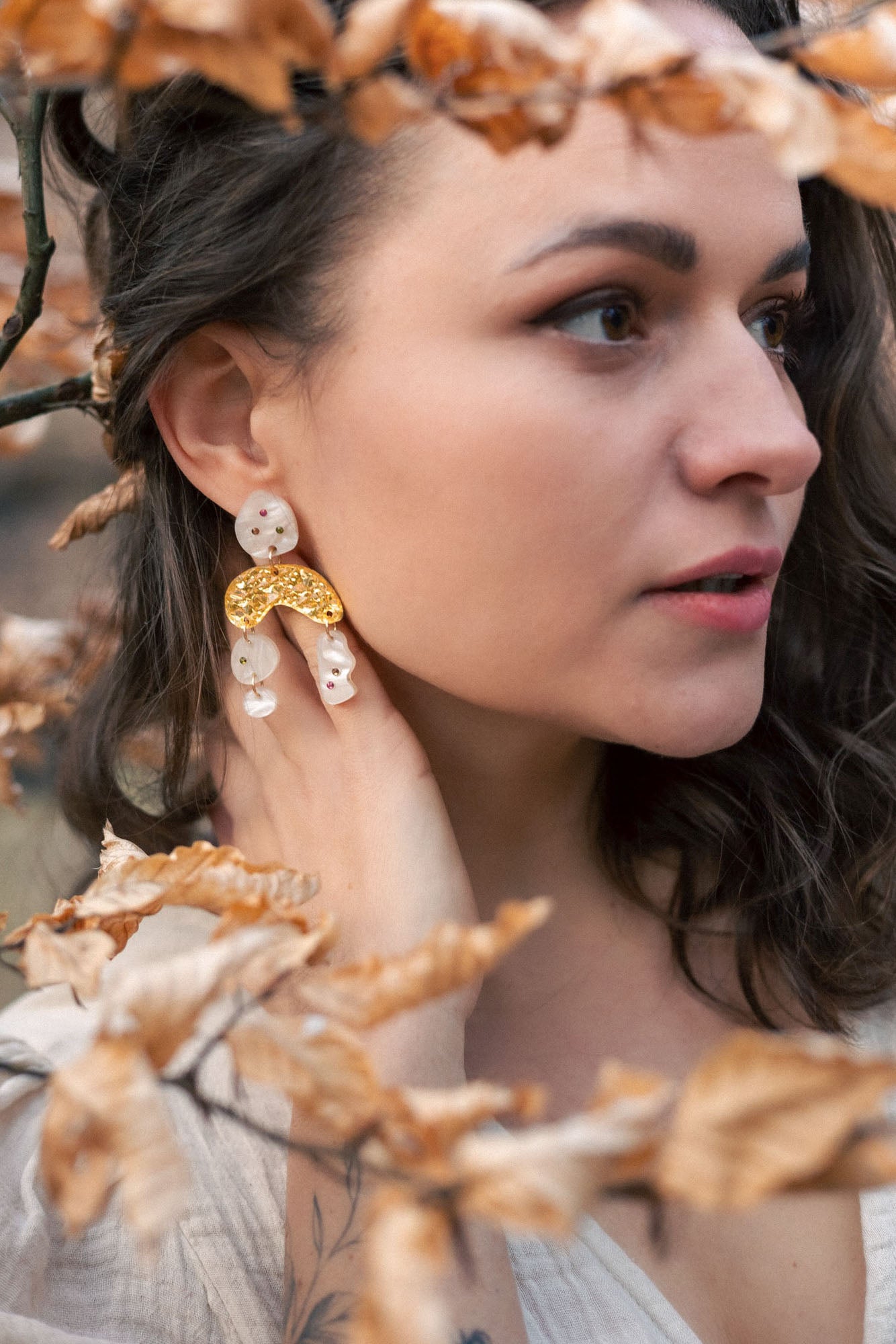 A woman with wavy brown hair wearing a cream top and boho chic dangle earrings stands among branches with dry, light brown leaves, gazing thoughtfully to the right.