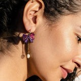 Close-up of a woman's ear with curly hair, wearing a gold ear cuff and an interchangeable dangle earring shaped like a purple bow with a dangling pearl. The side profile highlights her smooth skin and natural makeup.