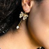 Close-up of a person's ear adorned with a gold-plated butterfly earring featuring a dangling pearl, reminiscent of elegant freshwater pearl earrings. The person has curly dark hair and a second small stud in the upper ear cartilage.