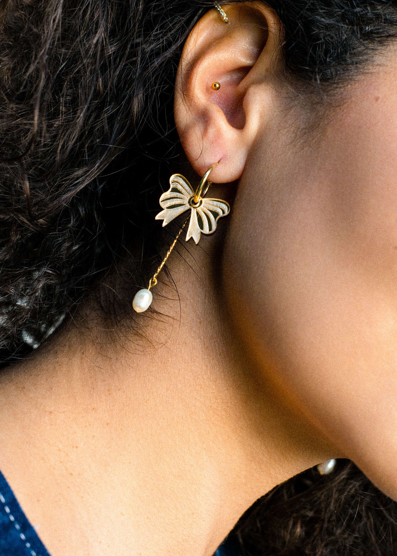 Close-up of a person's ear adorned with a gold-plated butterfly earring featuring a dangling pearl, reminiscent of elegant freshwater pearl earrings. The person has curly dark hair and a second small stud in the upper ear cartilage.