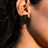 Close-up of a person’s ear with two earrings: a small gold stud in the upper ear and a large black circular earring with a gold accent in the earlobe. The black earring, crafted in an acrylic glass finish, adds a modern touch to the look. The person has curly dark hair.