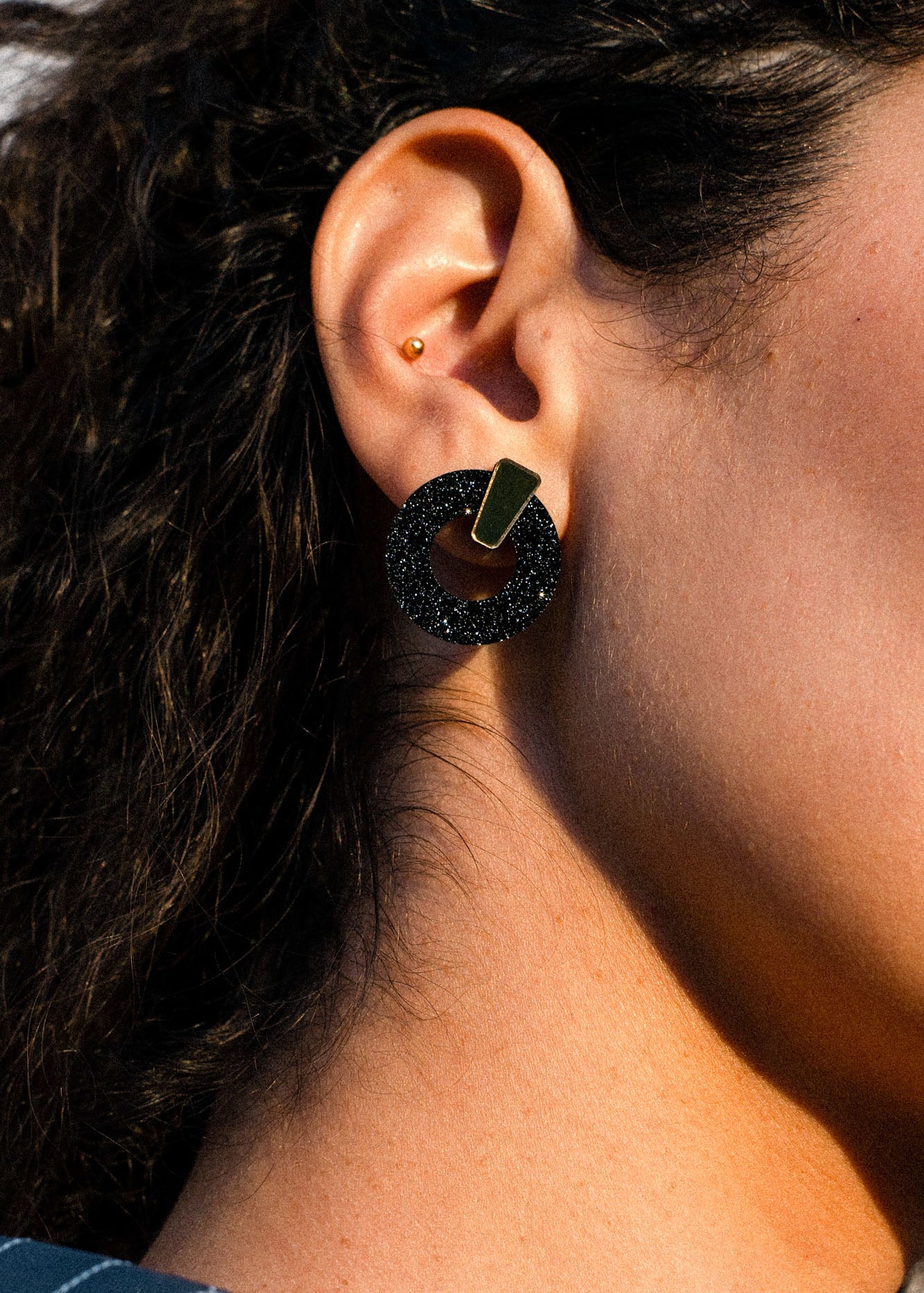 Close-up of a person’s ear with two earrings: a small gold stud in the upper ear and a large black circular earring with a gold accent in the earlobe. The black earring, crafted in an acrylic glass finish, adds a modern touch to the look. The person has curly dark hair.