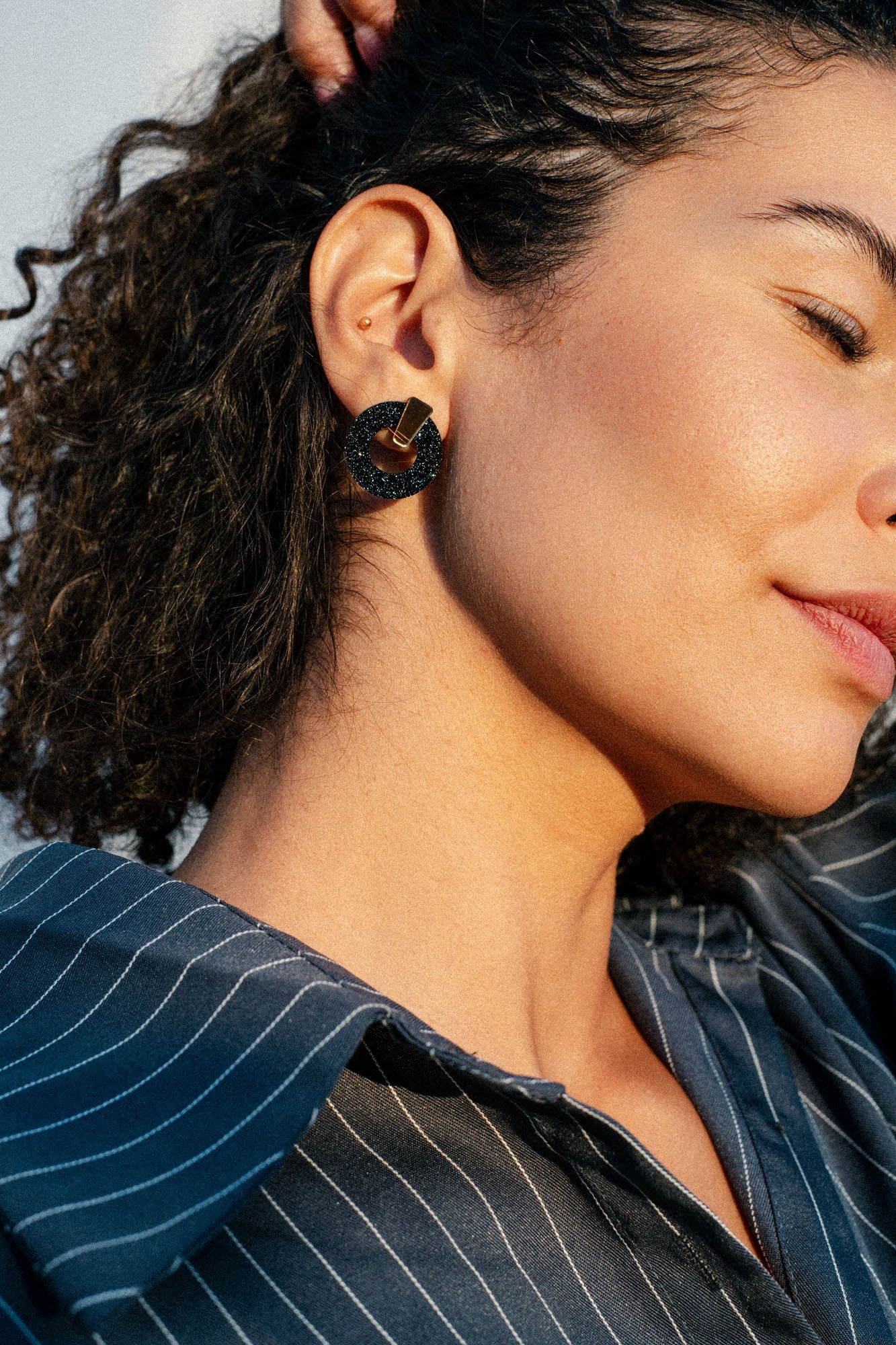 A woman with curly hair wears a dark, geometric acrylic glass earring and a pinstriped shirt. The image shows her profile and focuses on her ear and jawline, with sunlight highlighting her skin.