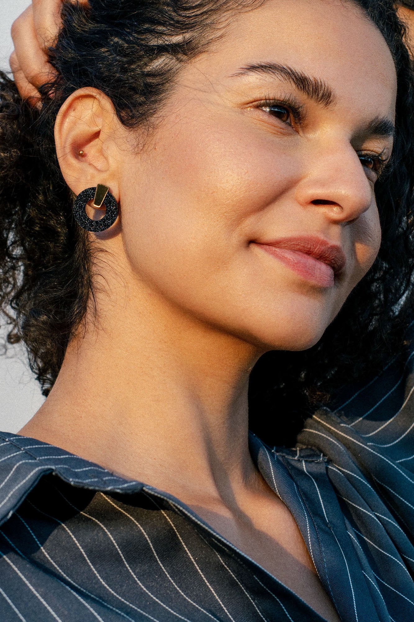 A woman with curly hair smiles gently, wearing a striped shirt and large round hypoallergenic earrings, with one hand behind her head, in soft natural light.