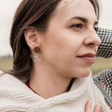 A woman in a houndstooth coat and cream scarf, embodying minimalist elegance, stands outdoors on a grassy, overcast day. She gazes thoughtfully into the distance, one hand on her chest and the other behind her head.