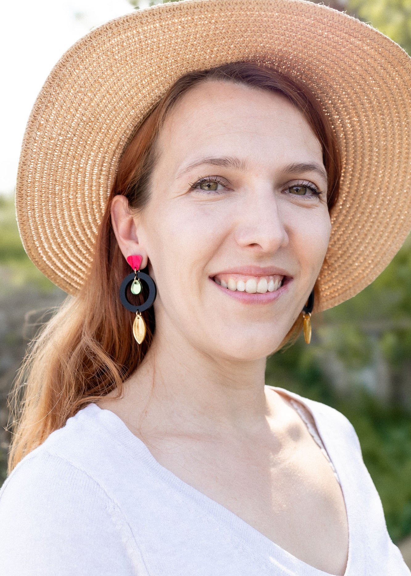A woman with light brown hair wearing a wide-brimmed straw hat, artisan dangle earrings, and a white top smiles outdoors, surrounded by greenery and sunlight.