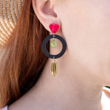 A woman in a straw hat and white shirt showcases artisan jewelry: dangle earrings crafted from recycled acrylic, featuring a black circle, gold shapes, and a red accent near the ear for a striking statement look.