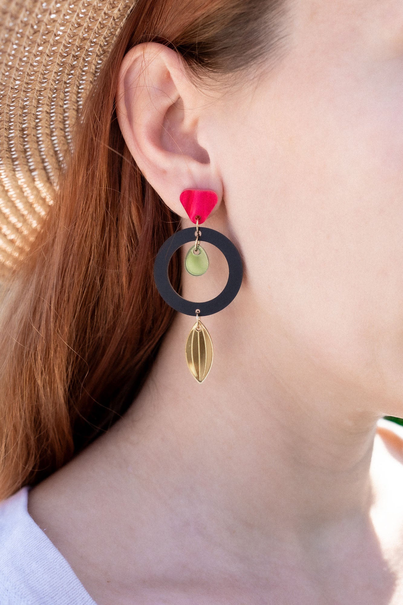 A woman in a straw hat and white shirt showcases artisan jewelry: dangle earrings crafted from recycled acrylic, featuring a black circle, gold shapes, and a red accent near the ear for a striking statement look.