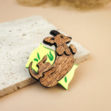 A playful brooch, this wooden pin shaped like a dachshund lounging on a yellow wedge with green leaf accents rests on textured beige stone, making it a charming dog accessory or a unique Dachshund brooch for any outfit.