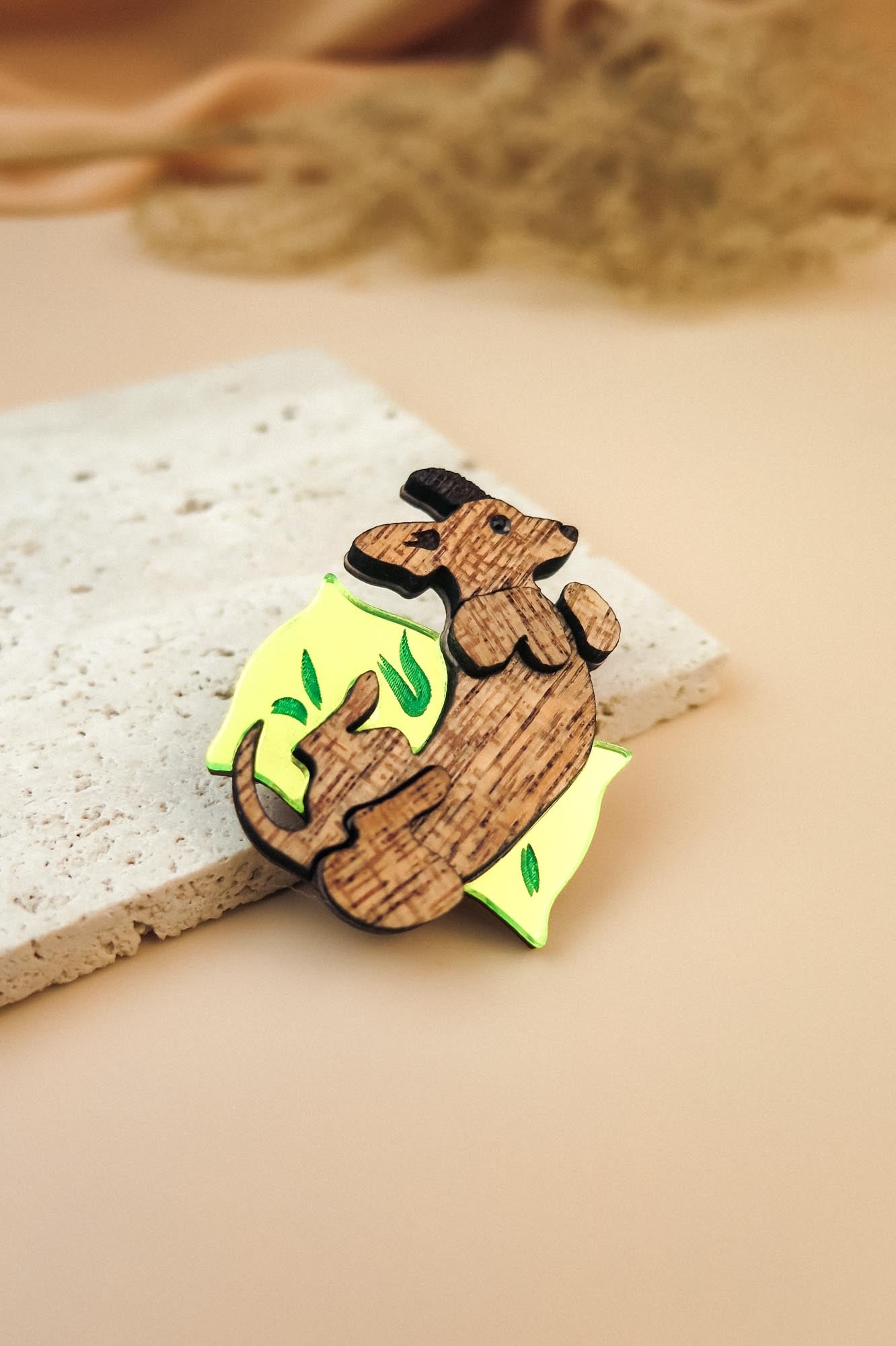 A playful brooch, this wooden pin shaped like a dachshund lounging on a yellow wedge with green leaf accents rests on textured beige stone, making it a charming dog accessory or a unique Dachshund brooch for any outfit.