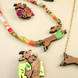 Assorted jewelry pieces featuring dachshund dog designs made of wood and colorful materials, including a playful brooch, necklaces, and unique dog accessories, displayed on a light beige surface.