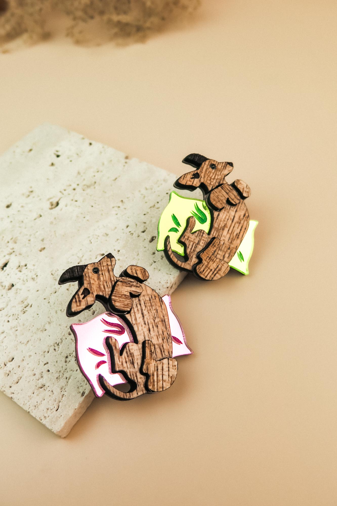 Two playful brooches shaped like reclining dachshunds, each resting on a colorful pillow—one pink and one neon green—placed on a textured stone surface. A charming touch for any dog accessory collection or fans of unique brooches.