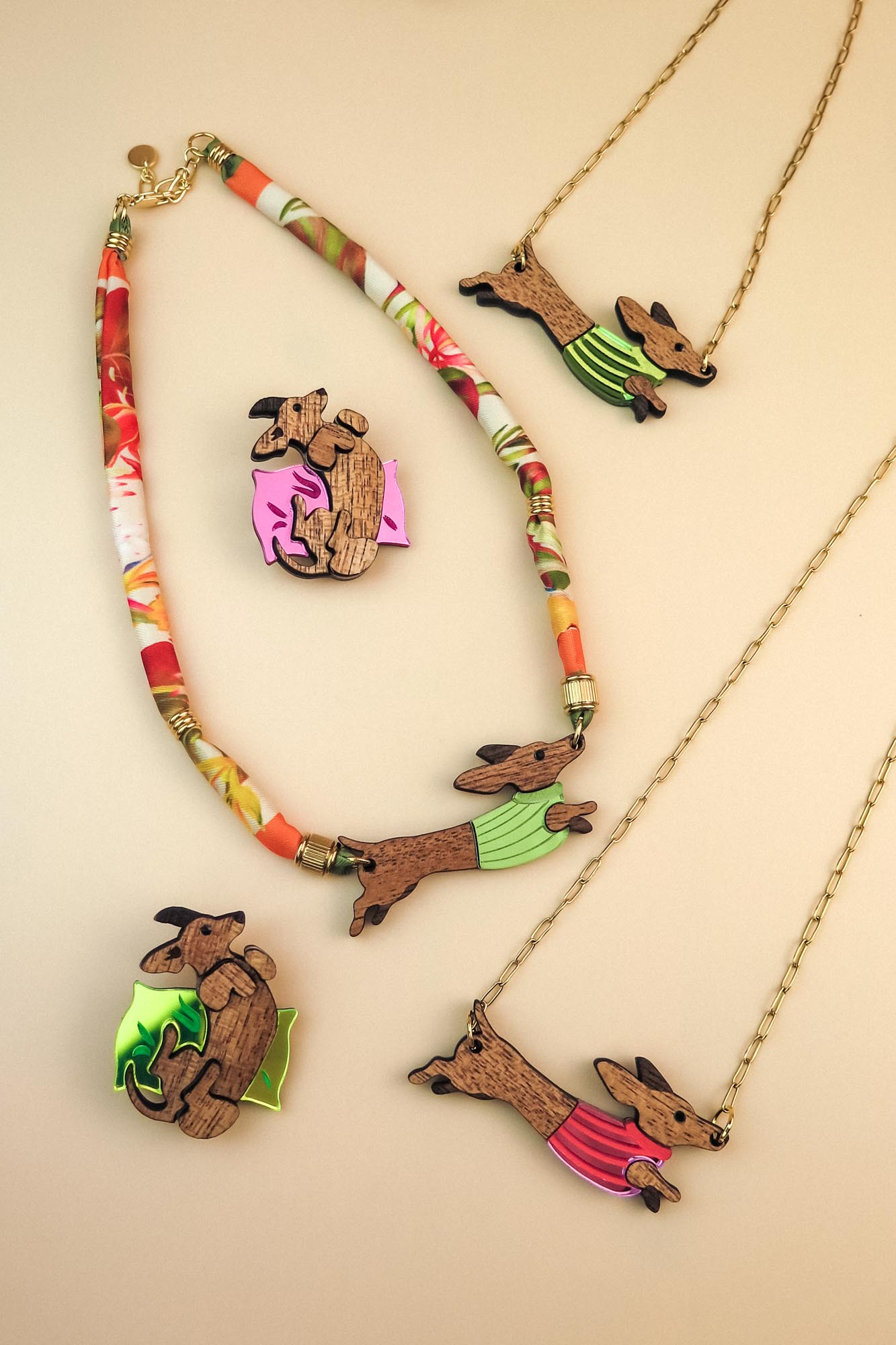 Colorful jewelry set featuring dachshund-shaped necklaces and brooches; the dogs wear green or pink colorful sweaters, with beads and gold chain accents, crafted from sustainable wood and arranged on a neutral beige background.
