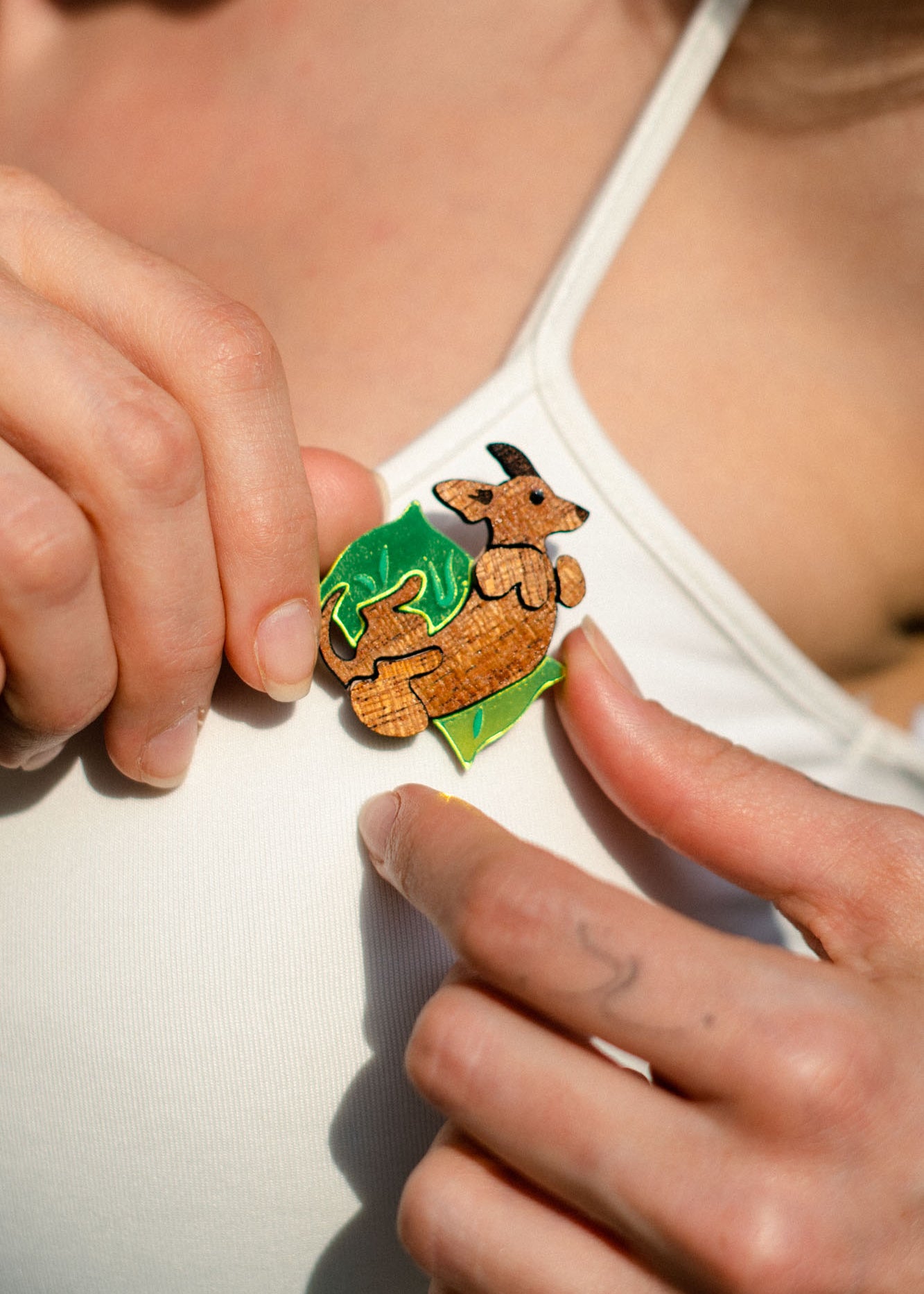 A person in a white tank top, surrounded by a green leaf backdrop, pins a playful brooch shaped like a kangaroo onto their shirt.