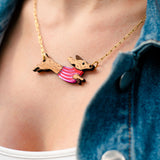 A close-up of a person wearing a gold necklace with a handcrafted Dachshund pendant in a pink striped sweater, made from sustainable wood. The person is also wearing a white top and a blue denim jacket.