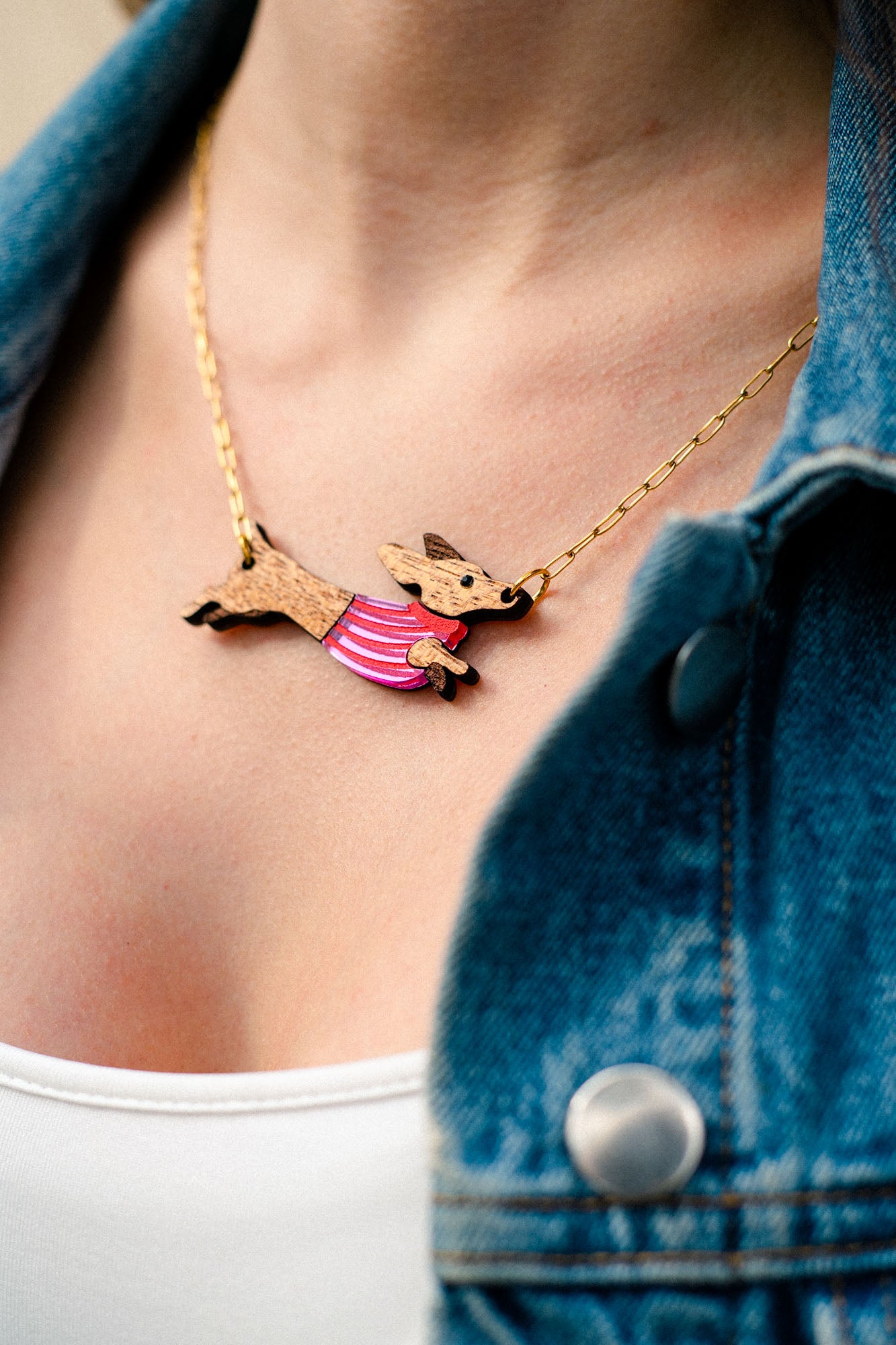 A close-up of a person wearing a gold necklace with a handcrafted Dachshund pendant in a pink striped sweater, made from sustainable wood. The person is also wearing a white top and a blue denim jacket.