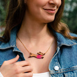 A woman wearing a denim jacket and white top smiles outdoors. Her long brown hair frames a colorful, handcrafted necklace featuring a Dachshund dog design made from sustainable wood.