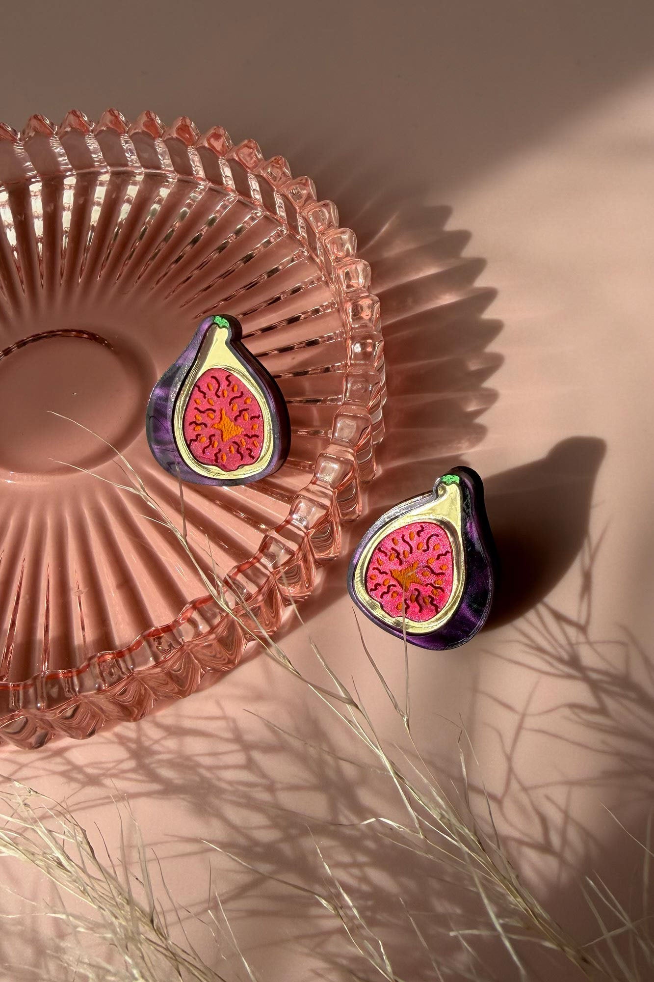 Two fig-shaped laser-cut studs with vibrant pink and deep purple detailing rest on a pink glass dish, casting shadows on a pale pink surface. Some delicate, dried grass lies nearby, adding texture to the scene.