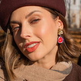 A woman wearing a burgundy beret and a beige scarf poses outdoors. She wears red lipstick, handcrafted fig earrings, and a matching pin. Autumn trees and a building create a scenic background.