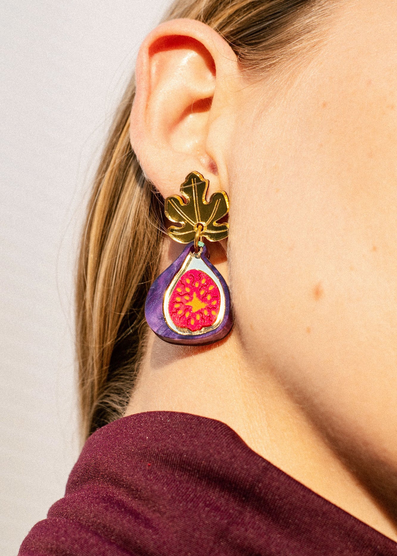 Close-up of a woman’s ear wearing handcrafted Fig Earrings shaped like a gold fig leaf with a colorful fig fruit design hanging below it. The woman has light skin, straight blonde hair, and the delicate earrings shimmer like gold mirror leaves.