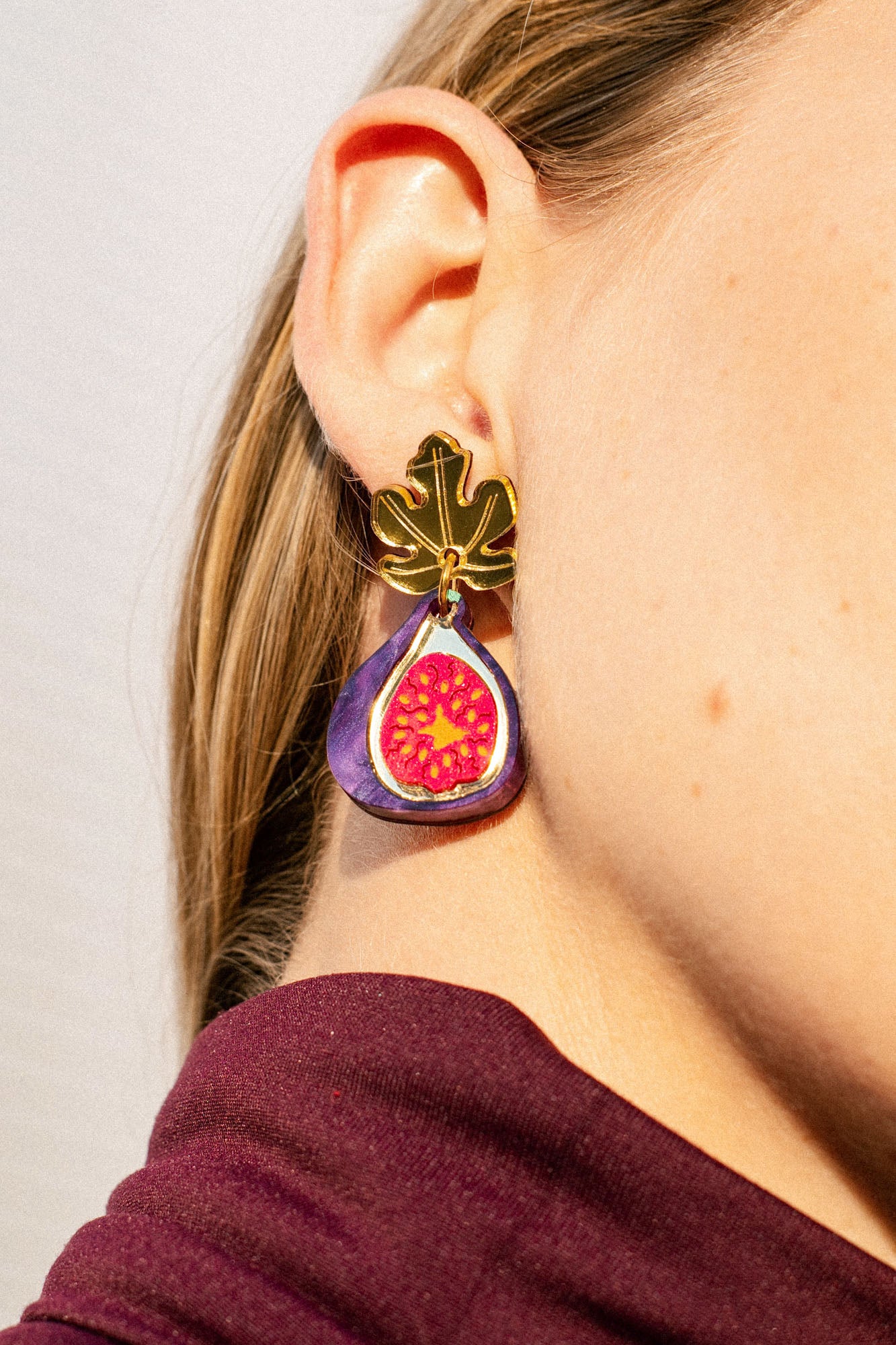 Close-up of a woman’s ear wearing handcrafted Fig Earrings shaped like a gold fig leaf with a colorful fig fruit design hanging below it. The woman has light skin, straight blonde hair, and the delicate earrings shimmer like gold mirror leaves.