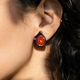 Close-up of a person’s ear wearing a colorful, hand-painted fig jewelry earring with purple and red details, against curly brown hair and sunlit skin.
