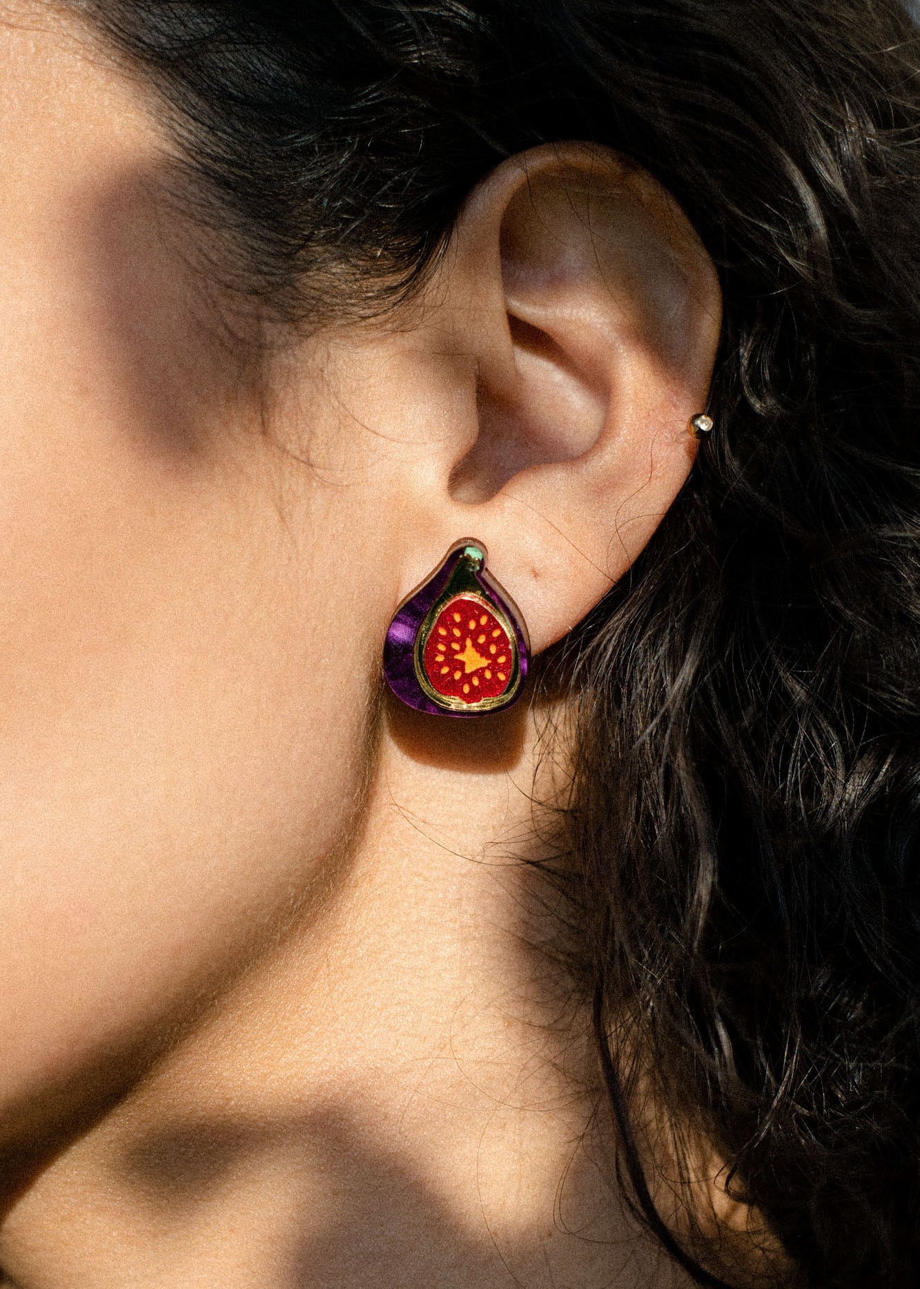 Close-up of a person’s ear wearing a colorful, hand-painted fig jewelry earring with purple and red details, against curly brown hair and sunlit skin.