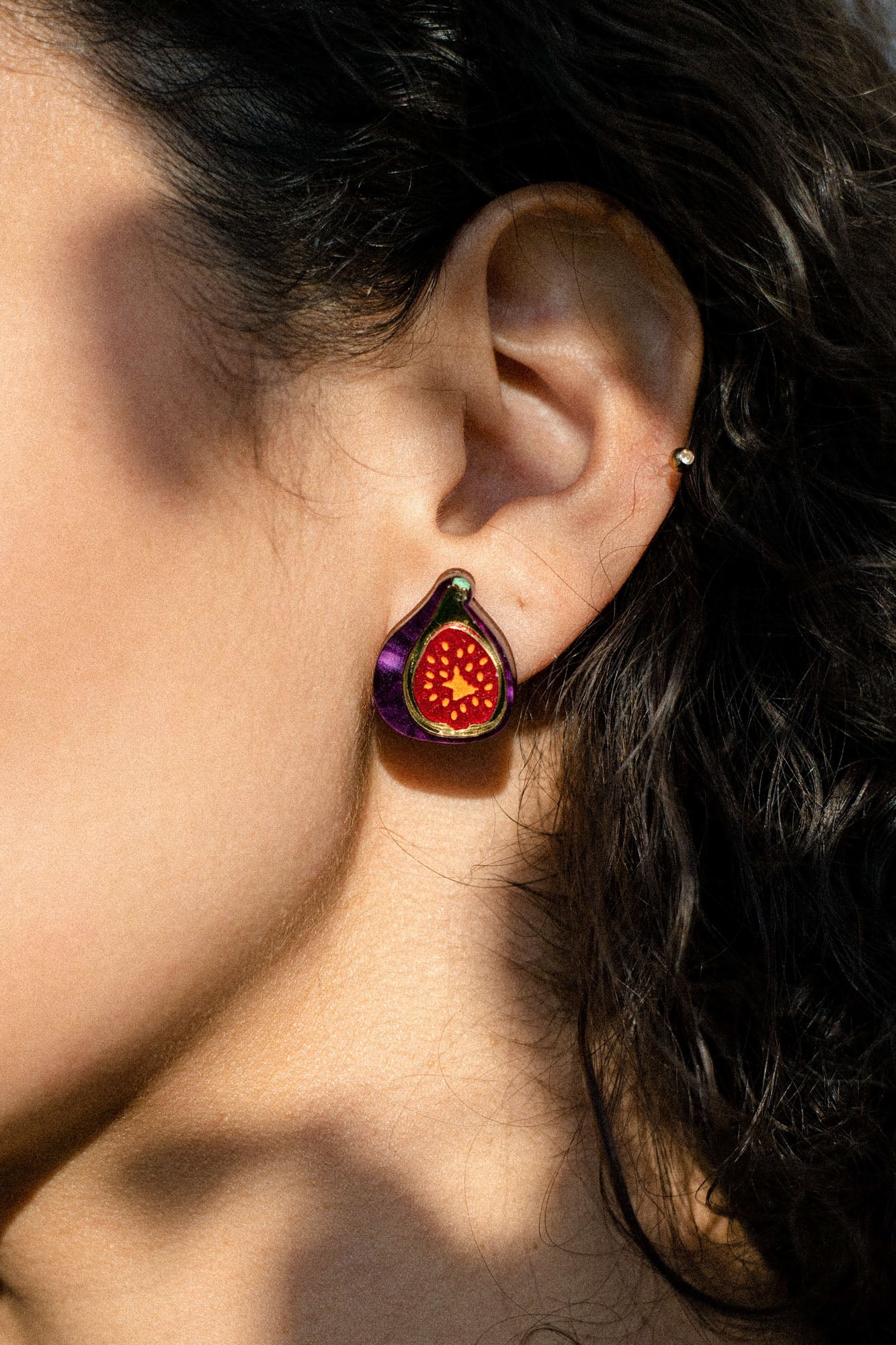 Close-up of a person’s ear wearing a colorful, hand-painted fig jewelry earring with purple and red details, against curly brown hair and sunlit skin.