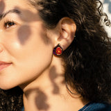 A woman with curly hair wears a hand-painted fig-shaped earring, standing in sunlight with dappled shadows on her face. Dressed in a blue top, she looks content, gazing slightly to the side.