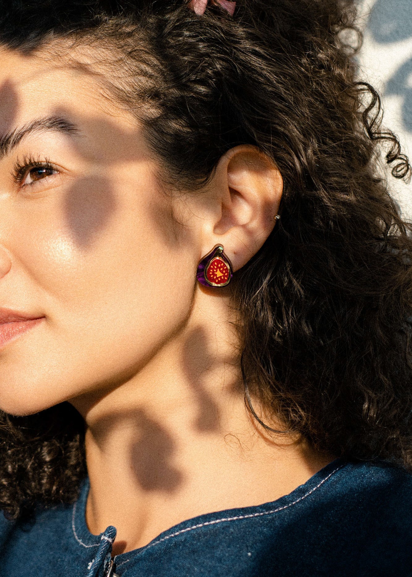 A woman with curly hair wears a hand-painted fig-shaped earring, standing in sunlight with dappled shadows on her face. Dressed in a blue top, she looks content, gazing slightly to the side.