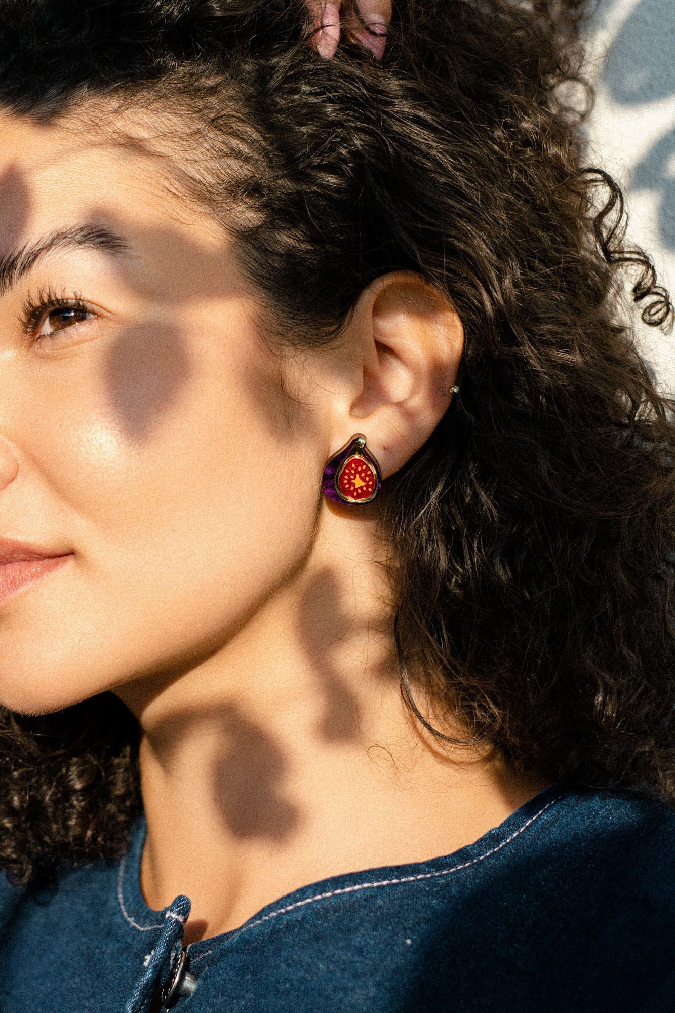 A woman with curly hair wears a hand-painted fig-shaped earring, standing in sunlight with dappled shadows on her face. Dressed in a blue top, she looks content, gazing slightly to the side.