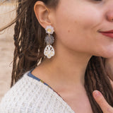 A woman with closed eyes and long brown dreadlocks wears a beige sweater and large handcrafted boho earrings, standing outdoors on a sandy beach. She smiles softly and touches her chest with one hand.