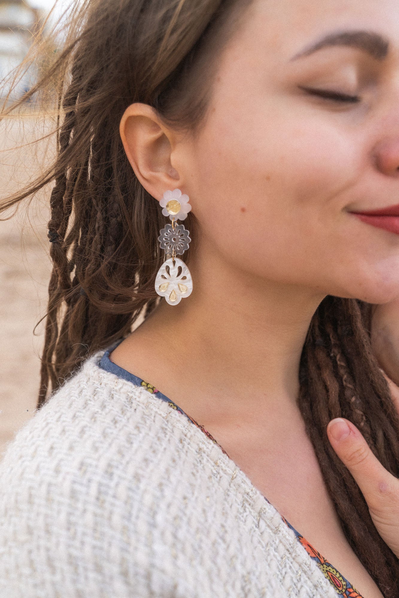 A woman with closed eyes and long brown dreadlocks wears a beige sweater and large handcrafted boho earrings, standing outdoors on a sandy beach. She smiles softly and touches her chest with one hand.