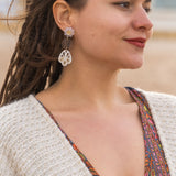 A woman with long braided hair stands outdoors, wearing a patterned dress and a cream knit sweater. She has handcrafted boho earrings and is smiling softly, looking to the side. The background appears to be a beach or sandy area.
