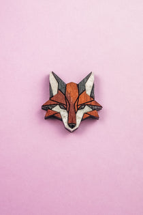 A geometric, stylized fox head brooch—handcrafted from recycled wood in shades of brown, black, and white—rests on a light pink background, making it an eco-conscious accessory with modern flair.