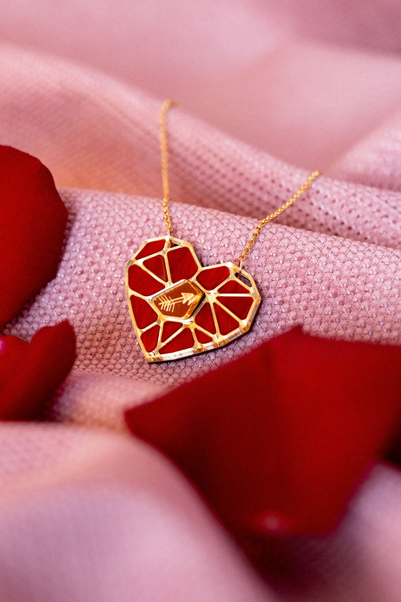 The handcrafted Fragment of Love Necklace features a heart pendant with a red, mosaic-like design, resting on soft pink fabric and surrounded by scattered red rose petals.