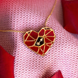 A handcrafted gold heart pendant, featuring a red mosaic design and a small white hand symbol, rests on pink textured fabric amid red rose petals—a striking Fragment of Love Necklace.